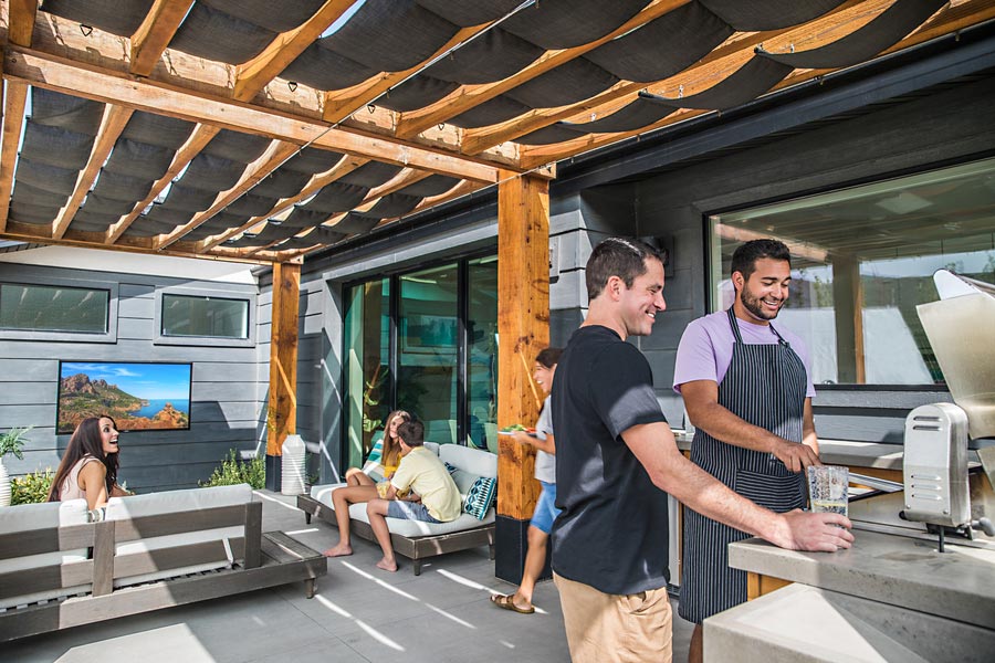 friends gather in an outdoor seating and grilling area. A wall-mounted Sunbrite TV shows an ultra-bright nature scene.