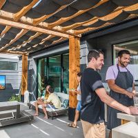 friends gather in an outdoor seating and grilling area. A wall-mounted Sunbrite TV shows an ultra-bright nature scene.