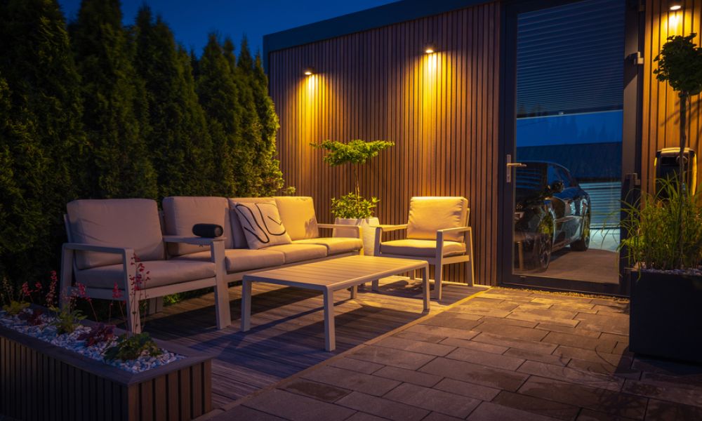 <span class="uk-h4">Downward-firing wall-wash light fixtures on the outside of a garage. Chairs and a portable speaker sit in front of the wall.</span>